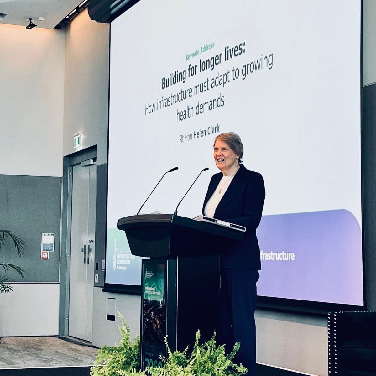 Helen Clark standing on stage in front of microphones and a screen at the Infrastructure Symposium 2026 organised by Te Waihanga | The New Zealand Infrastructure Commission