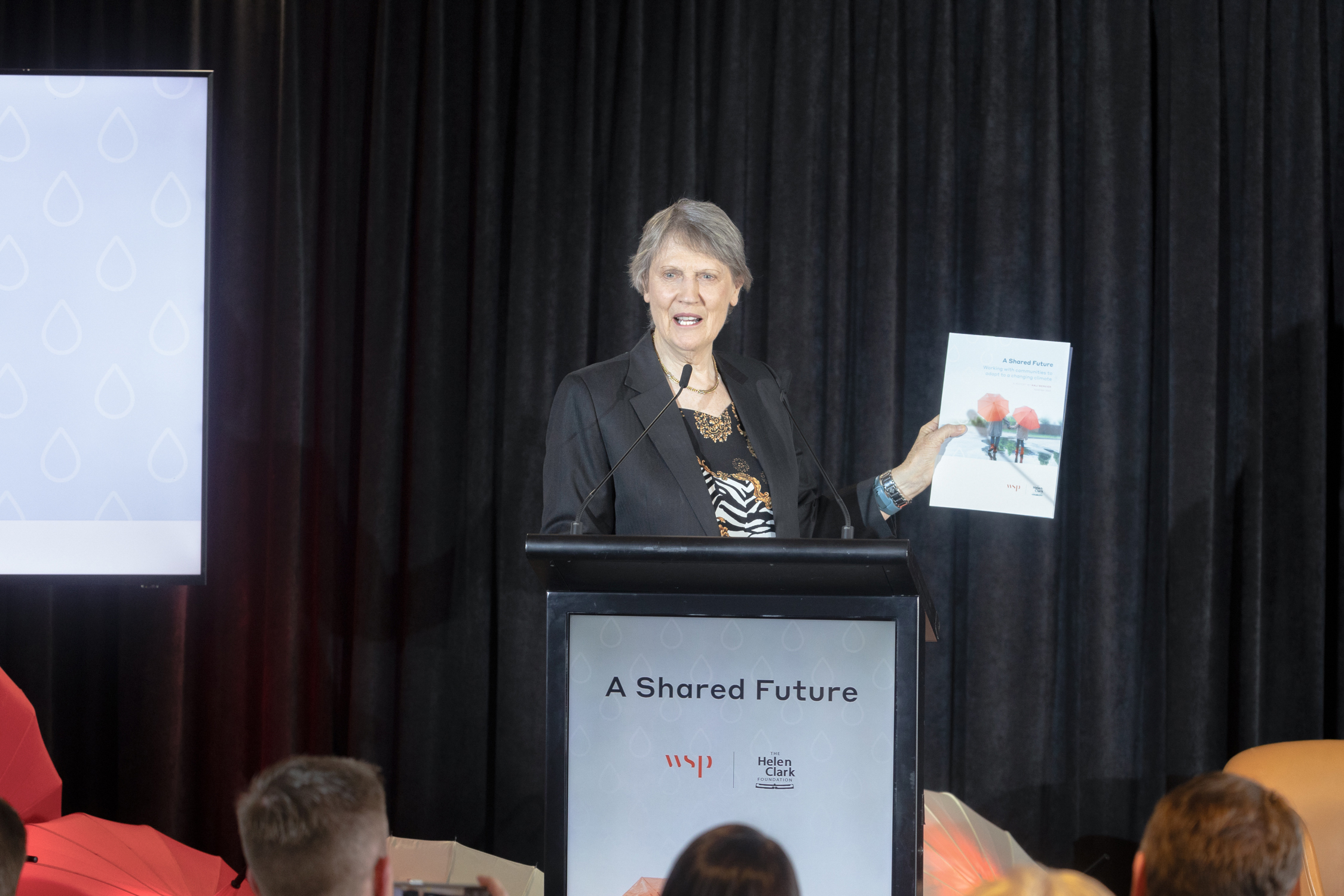 Helen Clark Foundation patron, Helen Clark, speaks at a podium holding a copy of the new report A Shared Future, which looks at involving communities in adaptation efforts and how meaningful community engagement can strengthen climate change adaptation.